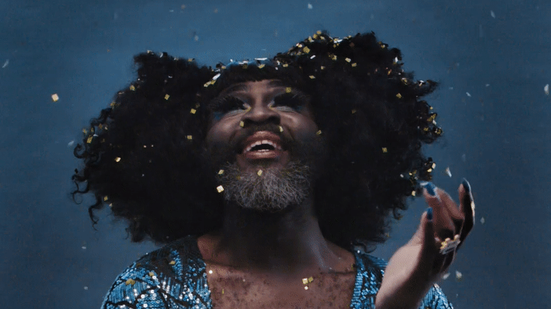 Twelfth Night Le Gateau Chocolat 2 wide A man in drag, wearing a blue sequinned dress, watches glitter fall from the sky around him.