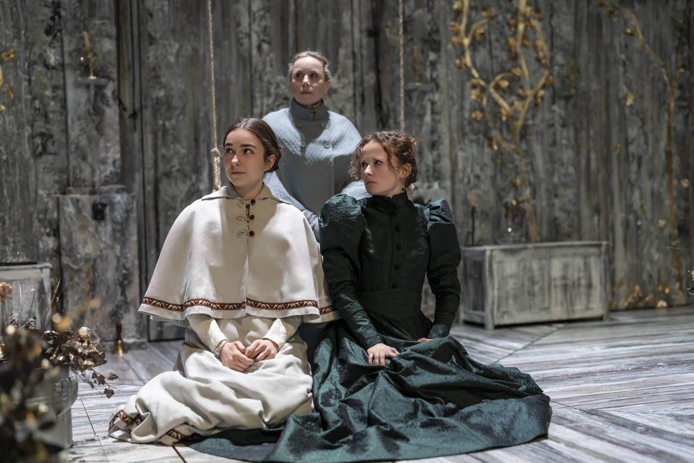 Ruby Thompson as Irina, Michelle Terry as Olga, and Shannon Tarbet as Masha in Three Sisters. Photographed by Johan Persson. Ruby Thompson as Irina, Michelle Terry as Olga, and Shannon Tarbet as Masha in Three Sisters. They are sat on the floor of the stage, wearing typical Russian costumes from the period, with elegant dresses and capes to keep out the cold.