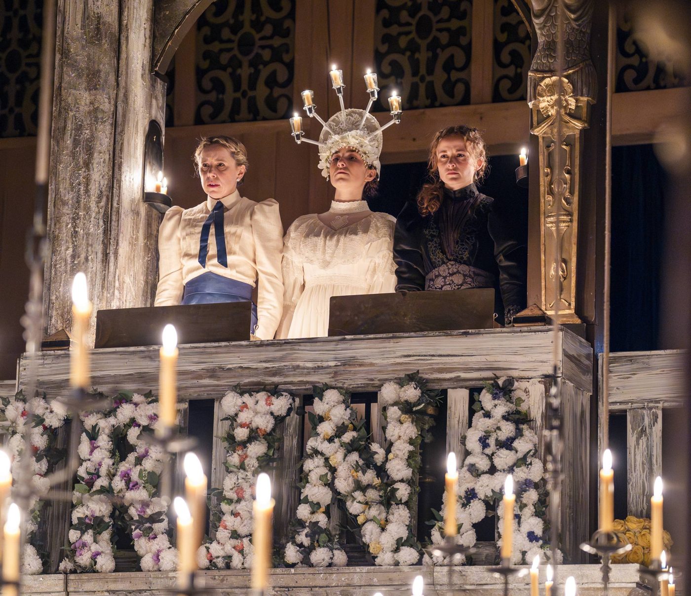 Michelle Terry as Olga, Shannon Tarbet as Masha and Ruby Thompson as Irina in Three Sisters. Photographed by Tristram Kenton. Michelle Terry as Olga, Shannon Tarbet as Masha and Ruby Thompson as Irina in Three Sisters. Olga wears a blouse and blue skirt, Irina wears a white lacey dress with a tiara made from lit candles, and Masha wears a dark black dress with her hair over one shoulder.