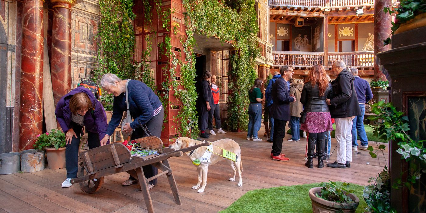 Access Ambassador Volunteer Shakespeare Globe Touch Tour PLM220917_113216 A group of people closely examine a garden set on a stage. With them is a guide dog.