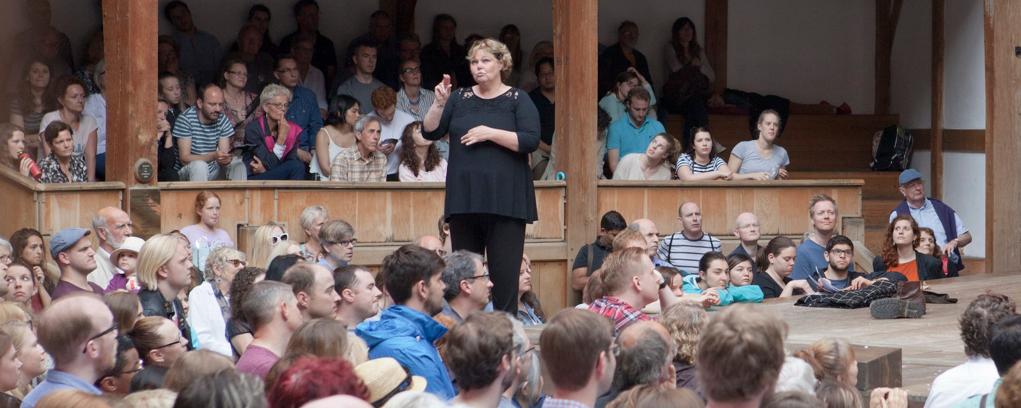 Access Performances British Sign Language Shakespeare’s Globe A woman stands on the outdoor wooden stage of the Globe Theatre and uses her hands to sign a performance to the audience