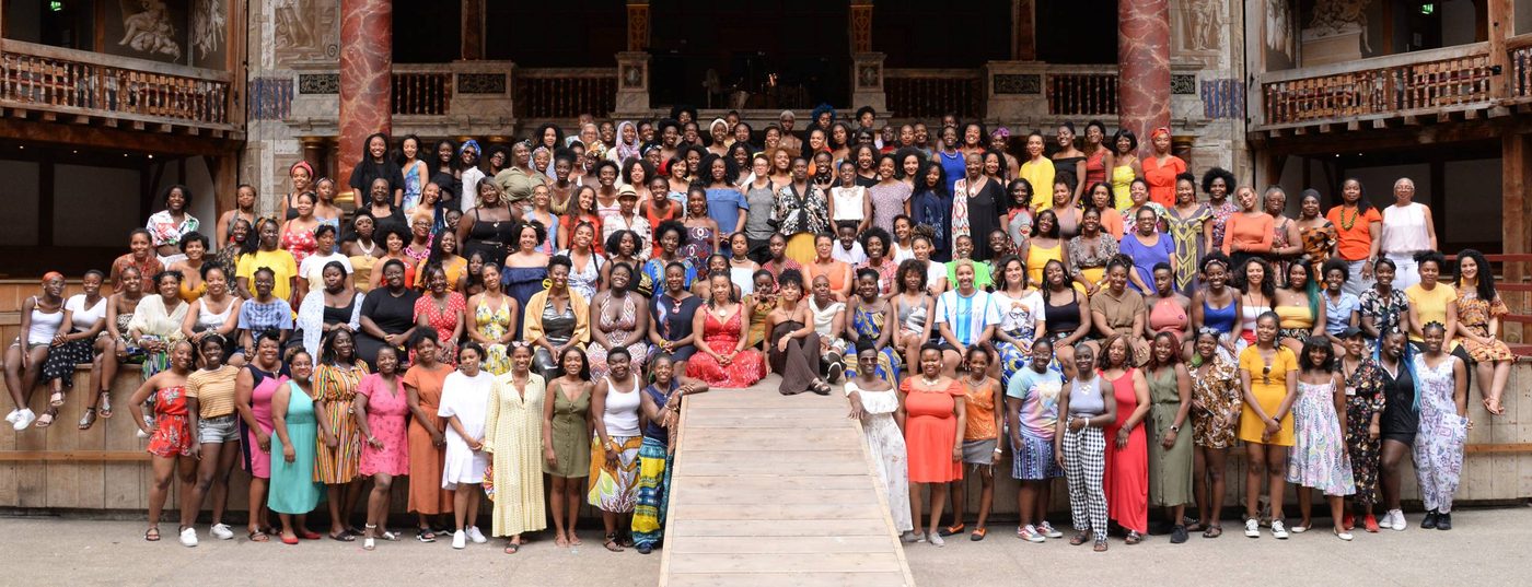 BLACK WOMEN IN THEATRE A group of people stand together on and around the stage of the Globe Theatre, smiling at the camera.