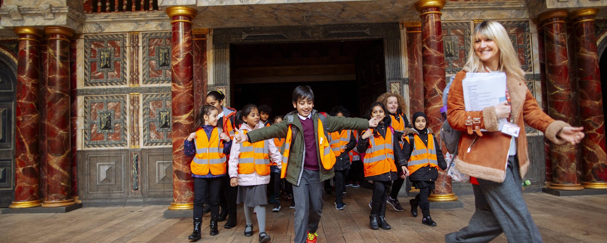 CD_6429_edit-scaled-aspect-ratio-2000-800 Group of primary school children in orange hi-vis vests following their teacher across the Globe stage