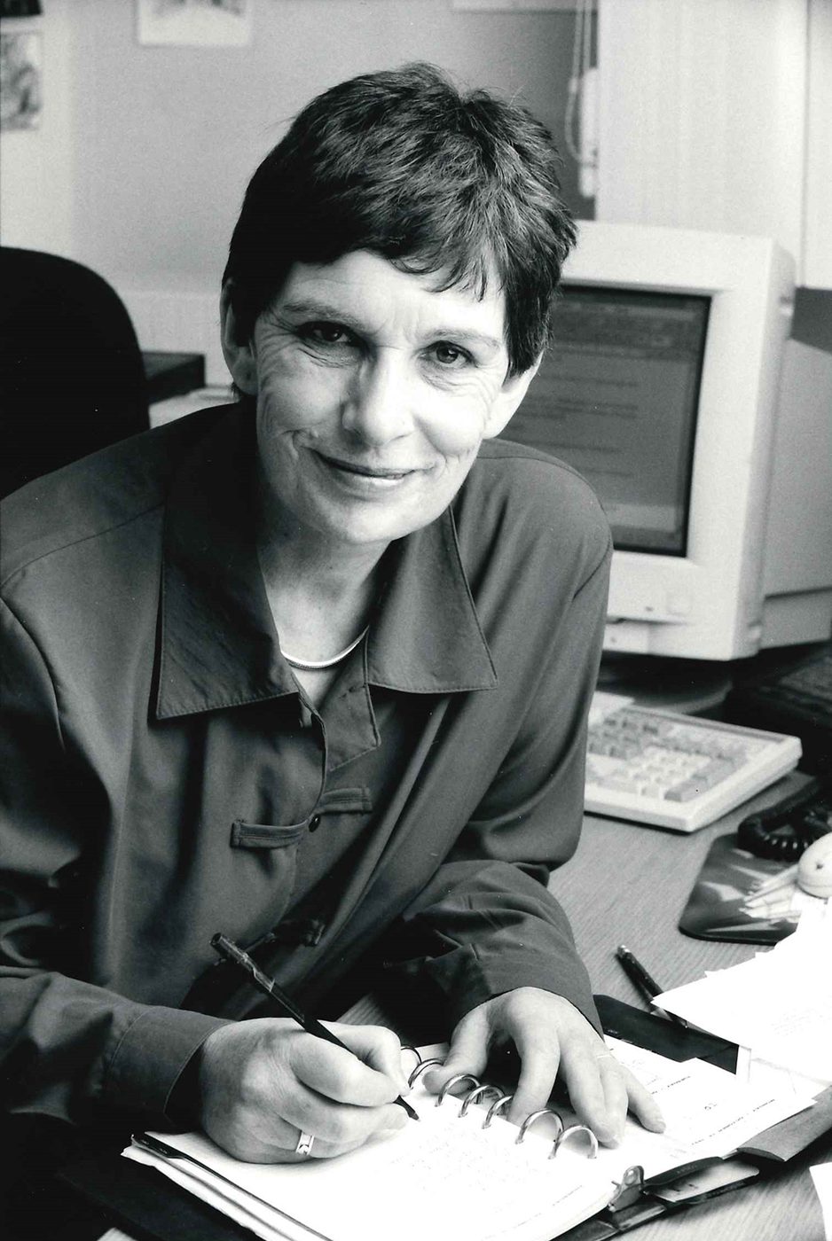 Globe Magazine Article Diana Devlin 1980s Portrait A black and white photograph of a woman smiling into the camera whilst writing in a notepad