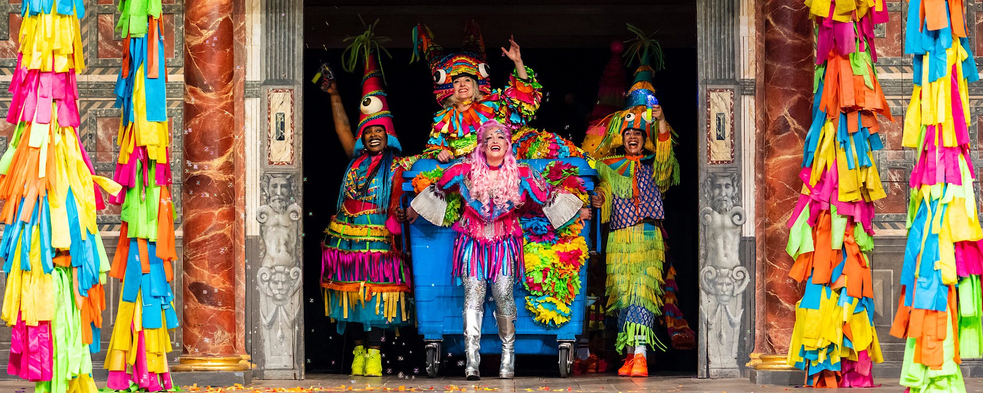 A Midsummer Night’s Dream 2021 Reviews Banner A group of actors bust onto the stage through doors, they are all dressed in bright colours and drag a blue wheelie bin which an actor sits on top of