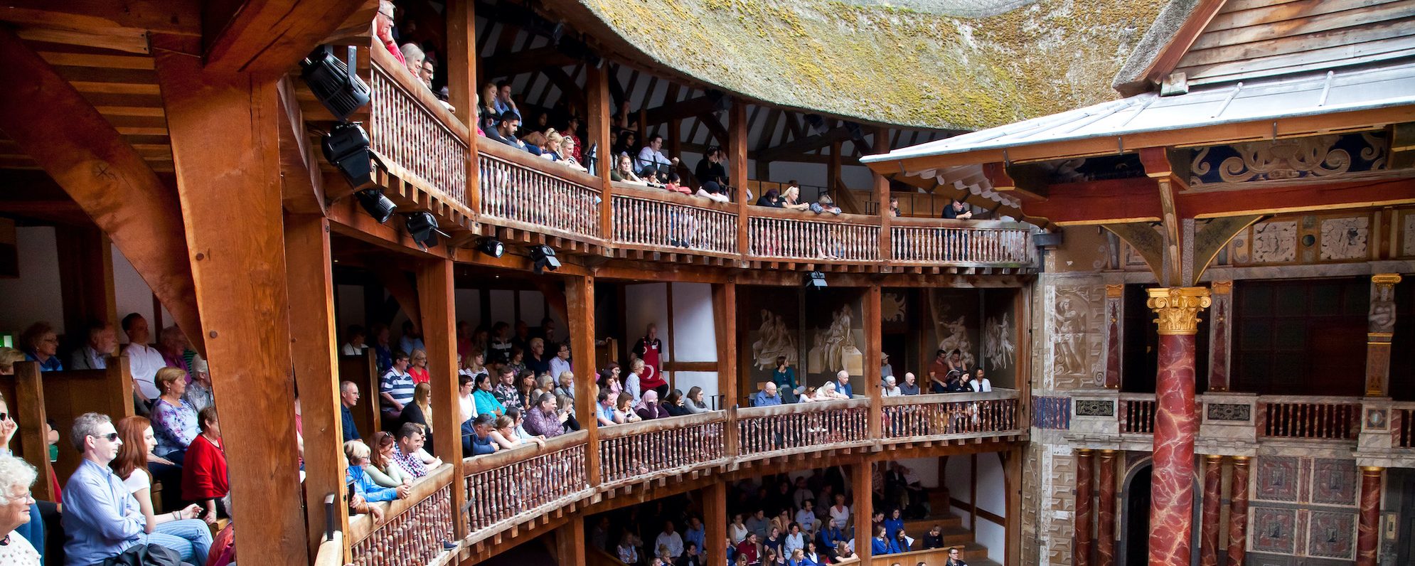 Globe Theatre Masthead Audience members sit on an outdoor balcony watching a play in the Globe Theatre