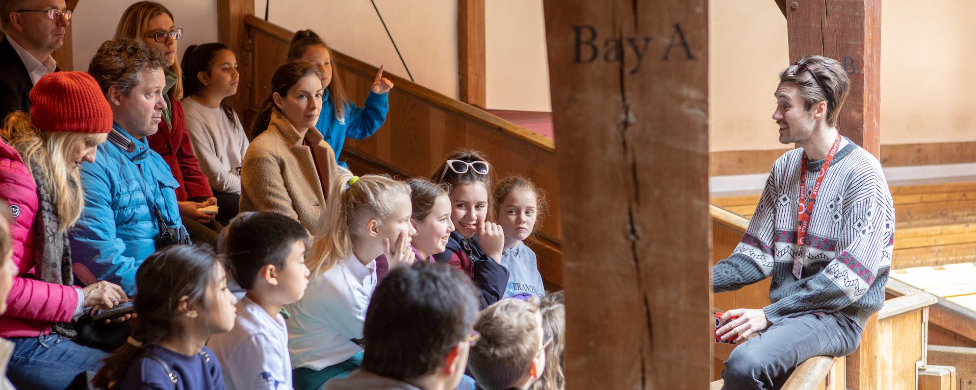 Plan Your Group Visit A guide talks to a group who are sitting on wooden benches in a theatre