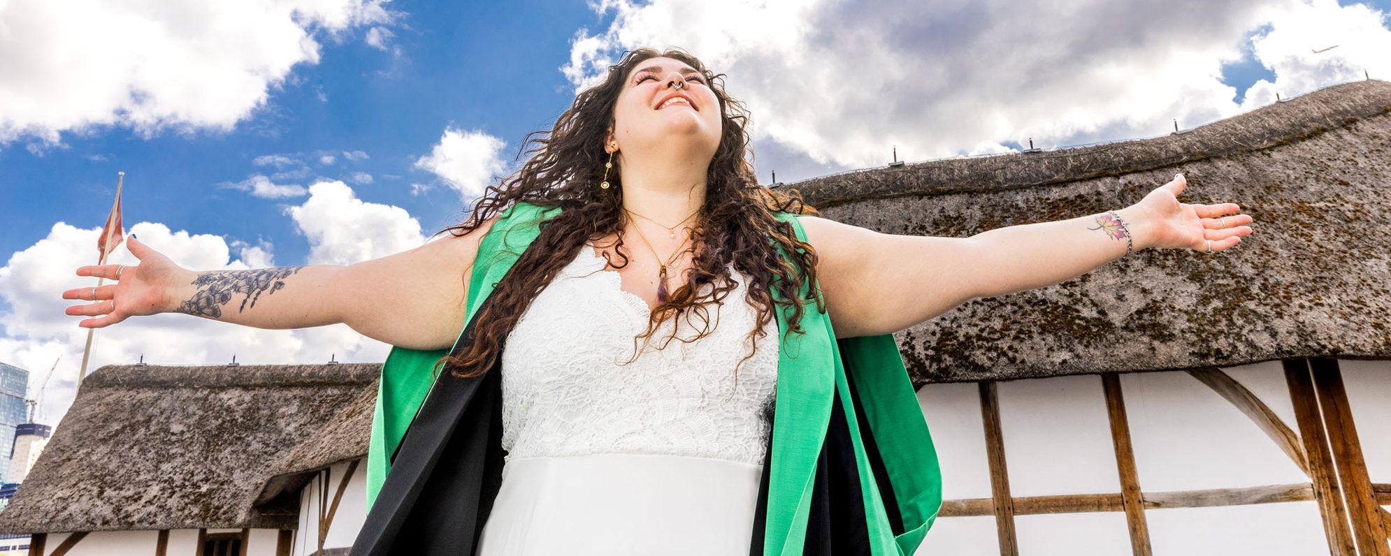 MA-Shakespeare-banner A woman wearing an academic graduation gown celebrates her MA with outstretched arms in front of the Globe.