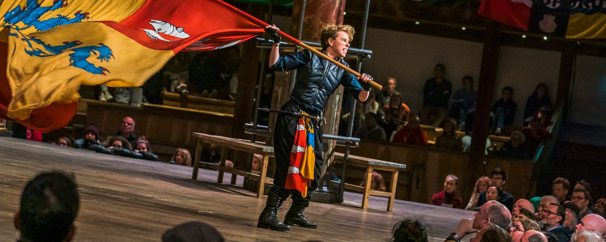 Press Room Masthead Henry V Part 1 Michelle Terry An actor on stage holding a large red and yellow flag