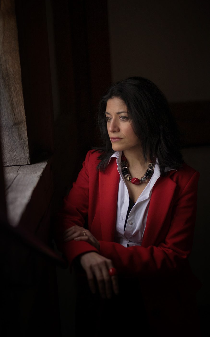 Professor Farah Karim-Cooper 8 Sarah Lee Web A woman wearing a red blazer looks out of a window, with daylight illuminating her.