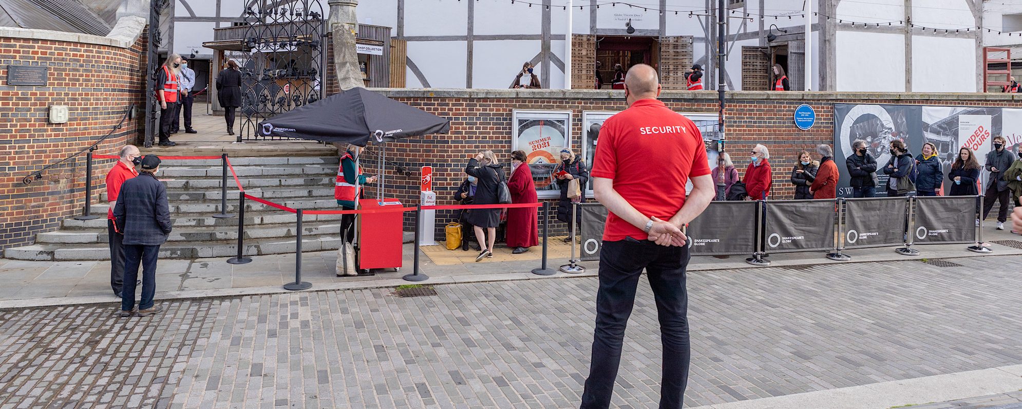 Security Visit Covid Coronavirus Shakespeare’s Globe Pete Le May 2021 A person with a red shirt that has the words ‘security’ written on the back of it stands in front of an outdoor white theatre