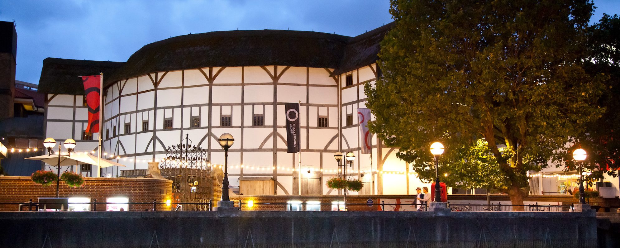 Shakespeare's Globe About Us Masthead Final A thatched theatre at night illuminated by string lights