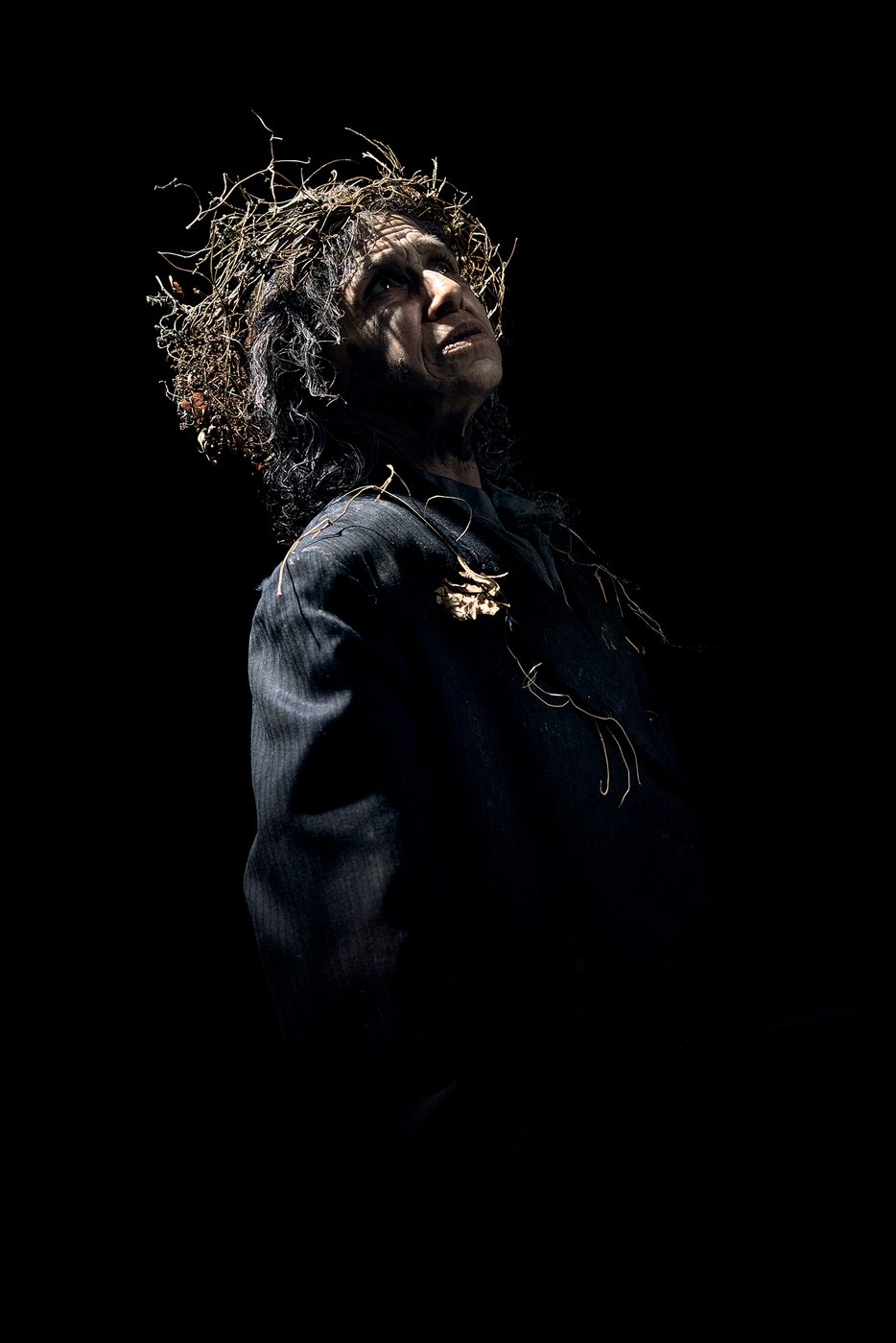 Shakespeares Globe_Kathryn Hunter_King Lear_Photographer Kate Bones_2022 An actor wearing a wreath crown of branches and leaves looks up, their face cast in shadow.