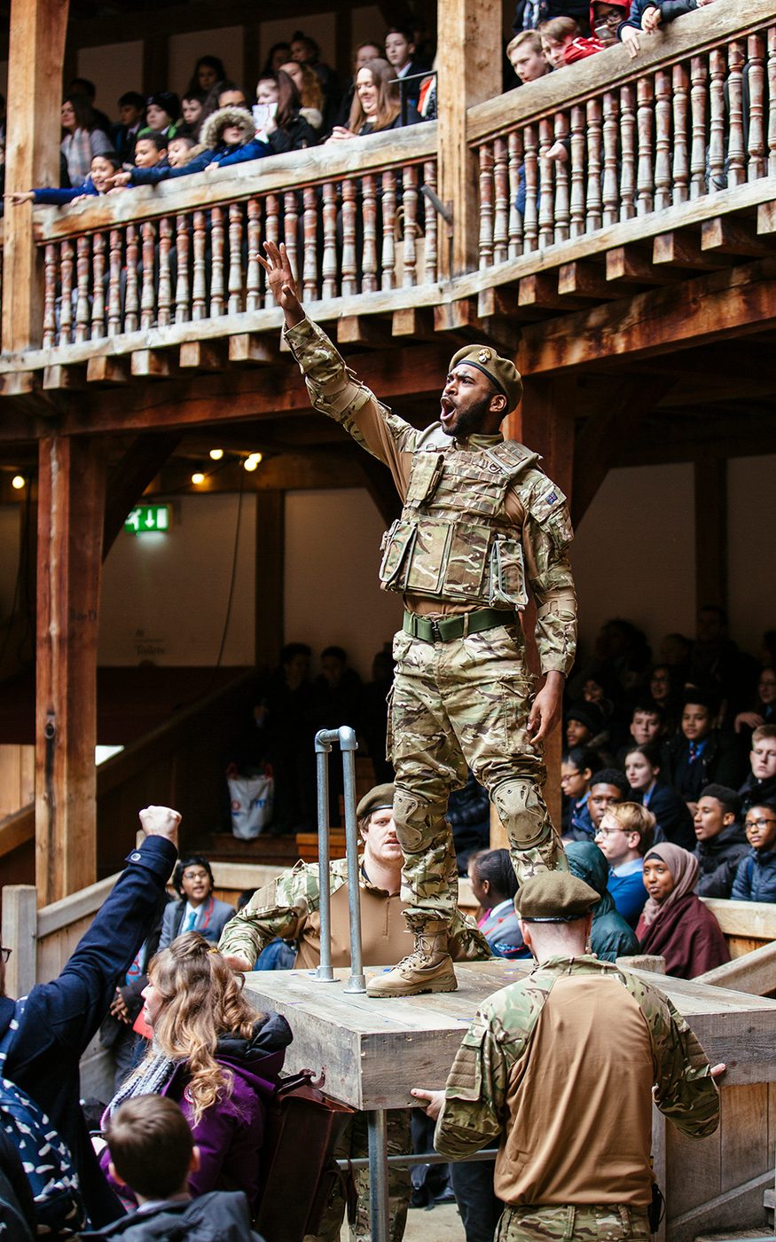Soldier Much Ado About Nothing 2018 Shakespeare's Globe Cesare De Giglio An actor dressed in modern military cameo uniform stands on a plinth in the Yard of the Globe Theatre, their arm outstretched pointing to the sky and shouting.