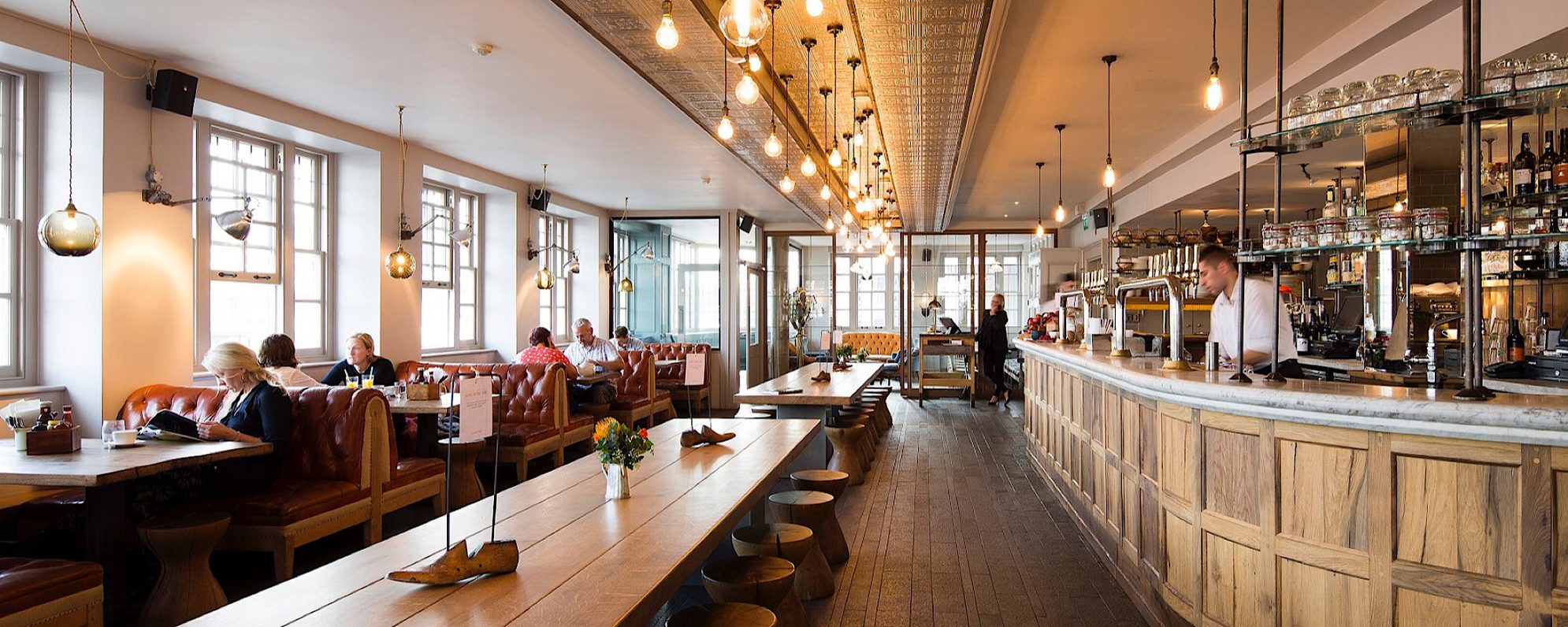 Swan Bar & Restaurant 2021 Shakespeare’s Globe Masthead A bright and open bar space has bench and booth seating, lit by large bulbs from the ceiling