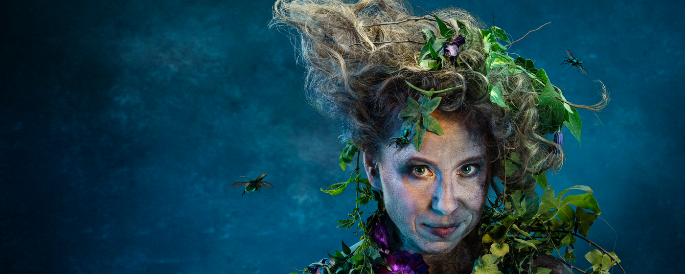 A Midsummer Nights Dream Study Days A close up of a smirking woman against a blue backdrop. She is covered in dirt and foliage and has insects flying around her unruly hair.