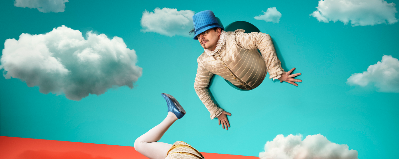The Comedy of Errors 2023 The torso of a man in Elizabethan dress appears from a hole in a blue backdrop with clouds. His legs appear from a different hole in the ground